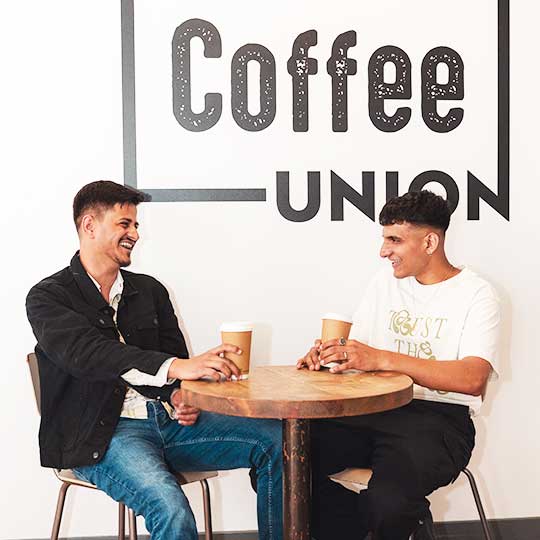 Two students sat in Coffee Union with the logo on the wall behind them