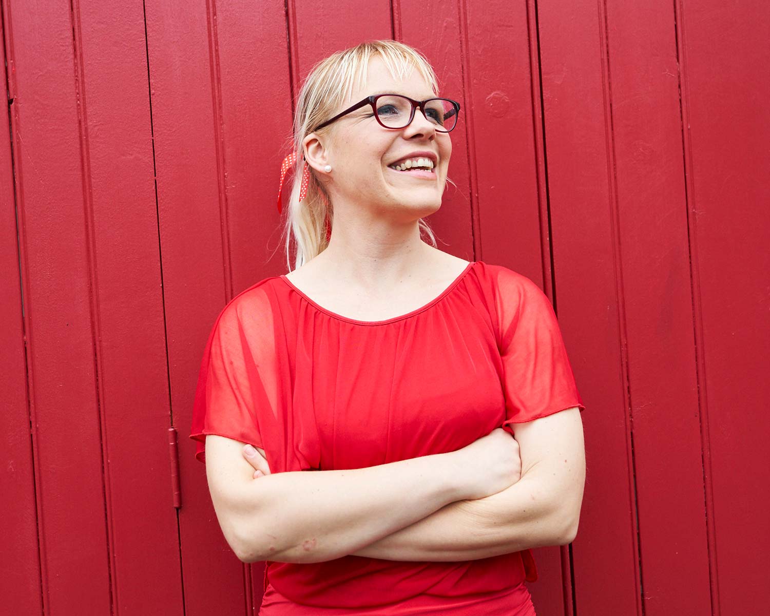 Tina stood in front of a red panelled door, her arms are folded and she's smiling at something off to the right of the camera